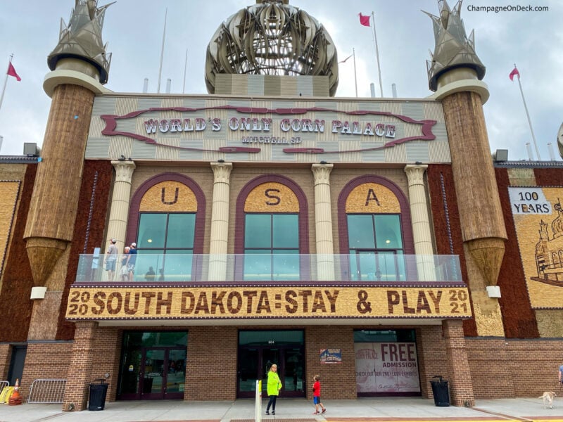 two kids stand outside of the exterior of the world's only corn palace, with three arched windows, turrets, and a banner than reads "2020 south dakota: stay and play 2021"