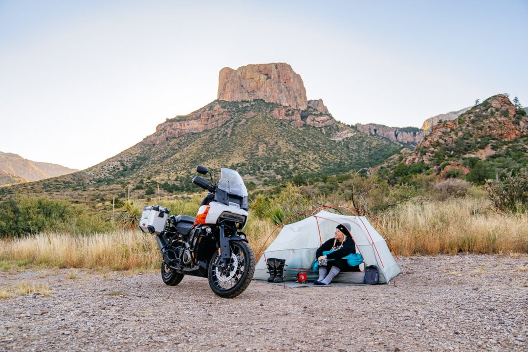 A woman sitting in a tent looking at a motorcycle parked next to it, surrounded by beautiful scenery
