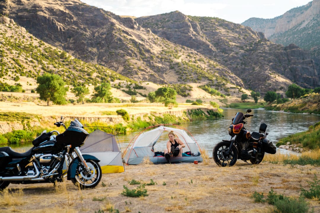 A woman sitting in a tent and taking her boots off, with a river and mountains behind her. Two motorcycles are parked next to her.