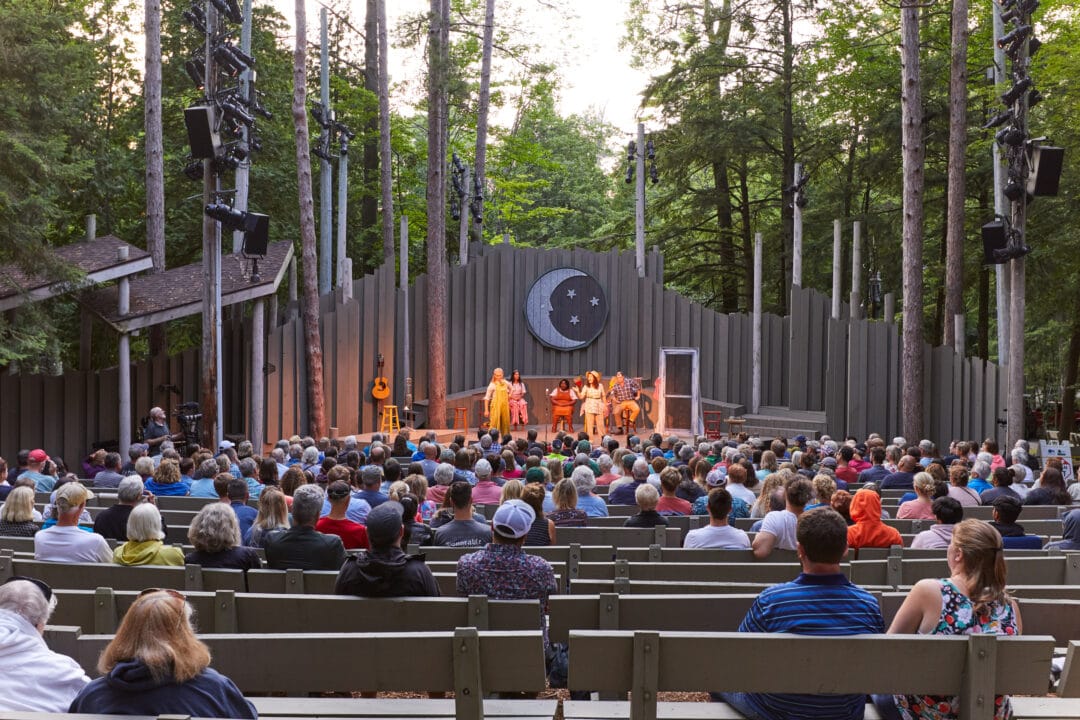 people sit on benches and watch a stage production at an outdoor ampitheater