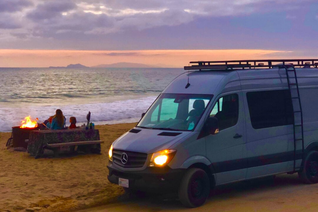 a white campervan is parked on the beach at sunset while people sit near a campfire