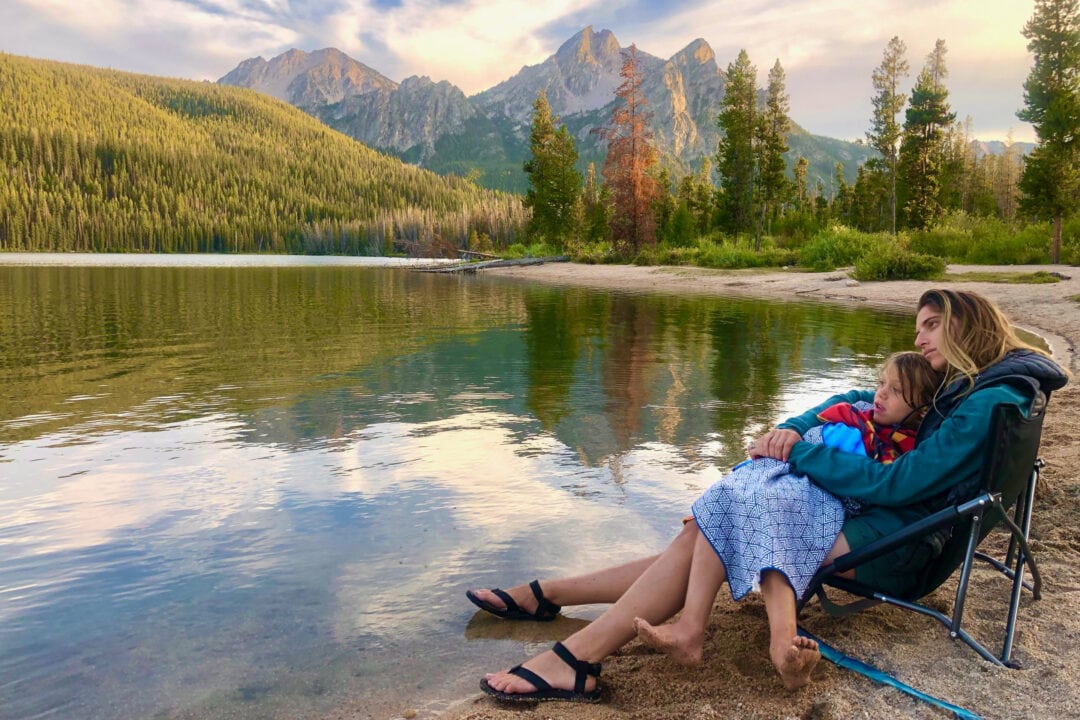 a woman and child sit in a chair next to a lake in front of mountains