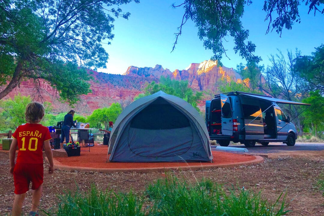 two people stand next to a white campervan parked at a campsite with a tent in front of red rocks