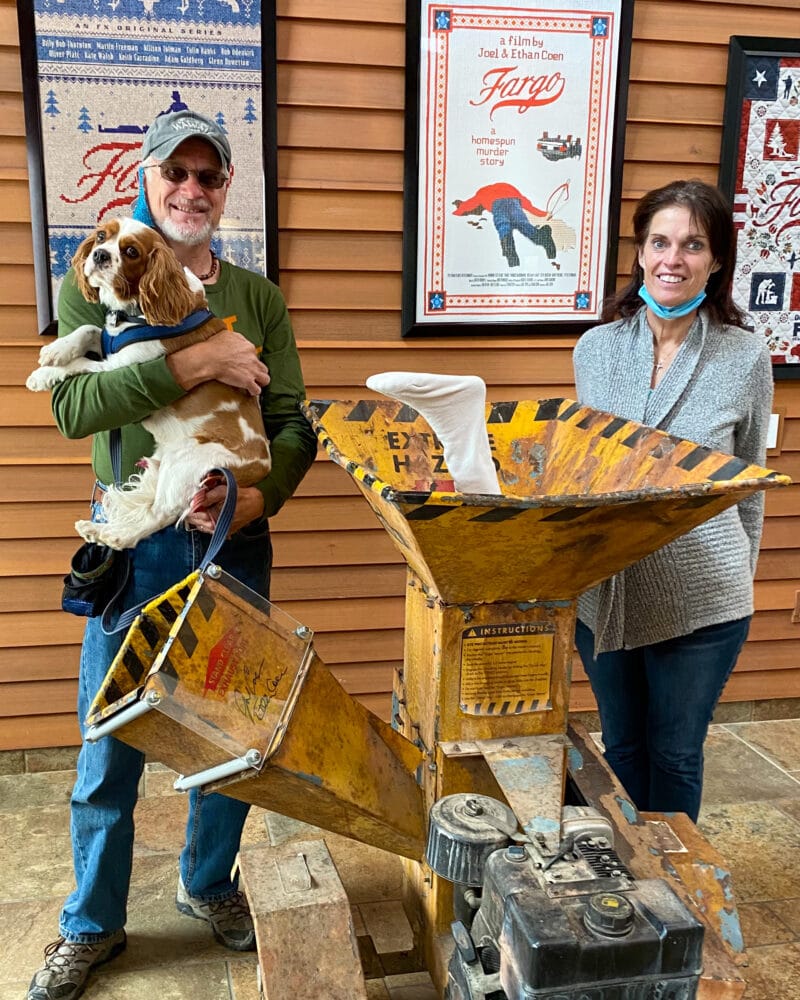 a man and woman and dog stand in front of a bright yellow wood chipper