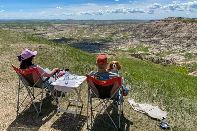 a couple and a dog sit in foldable camp chairs and take in a view of rock formations against a blue sky