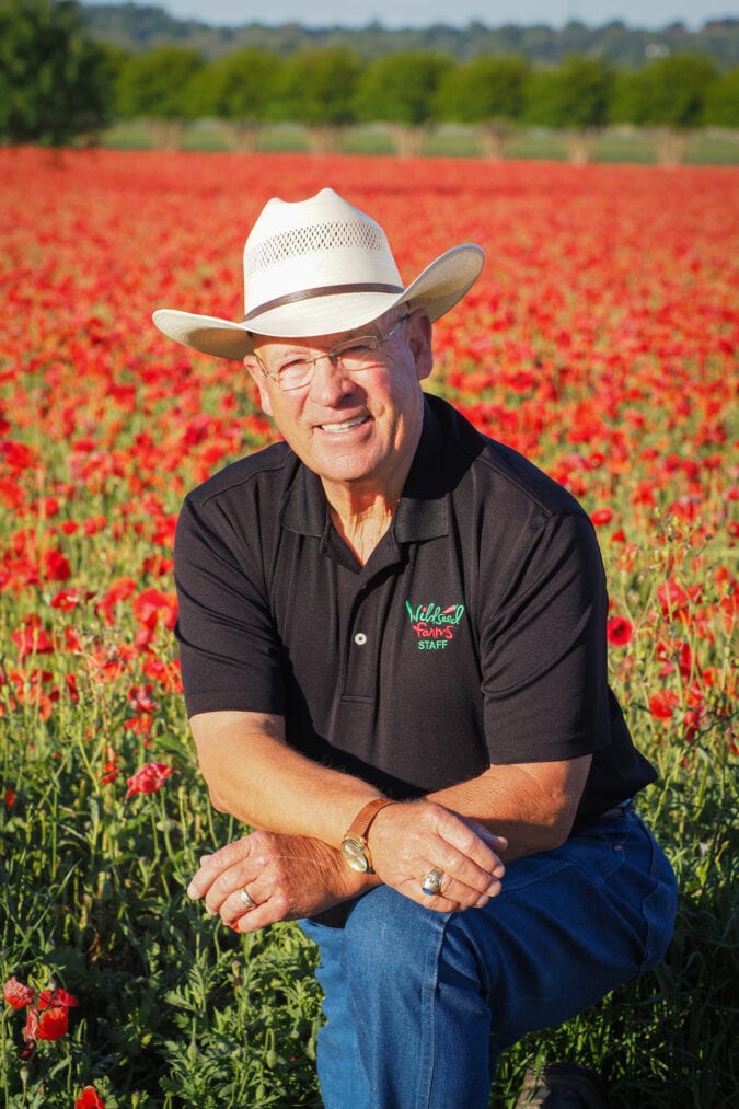 a man in a cowboy hat kneels in a field of bright red wildflowers
