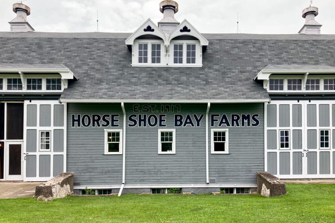 a grey barn with white trim with black lettering painted on the side that reads "est. 1917 horseshoe bay farms"