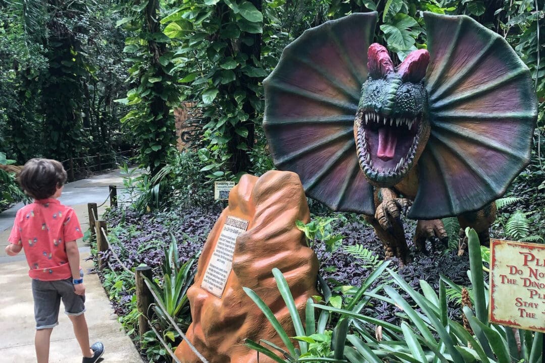 a boy walks by a dinosaur statue in an outdoor park