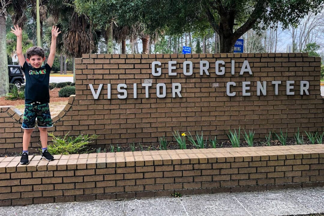 a boy stretches his arms above his head and poses next to a brick sign for the georgia visitor center