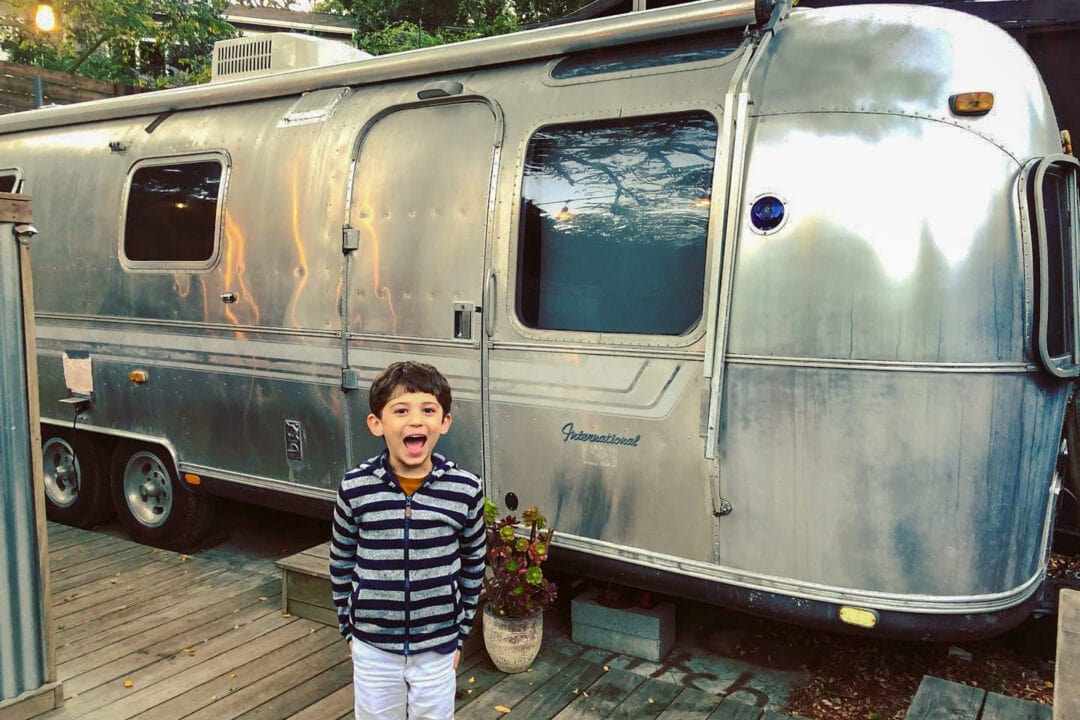 a boy in a striped shirt and white shorts stands in front of a silver airstream trailer
