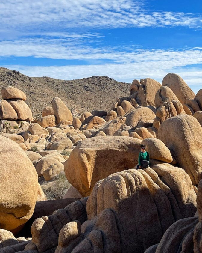 a boy sits on large rock formations against a blue sky in joshua tree national park
