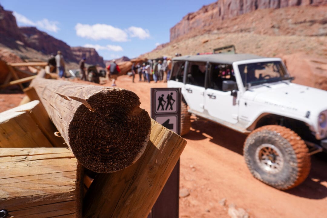 Close-up of fencing with a red rock desert landscape, a Jeep, and people in the background