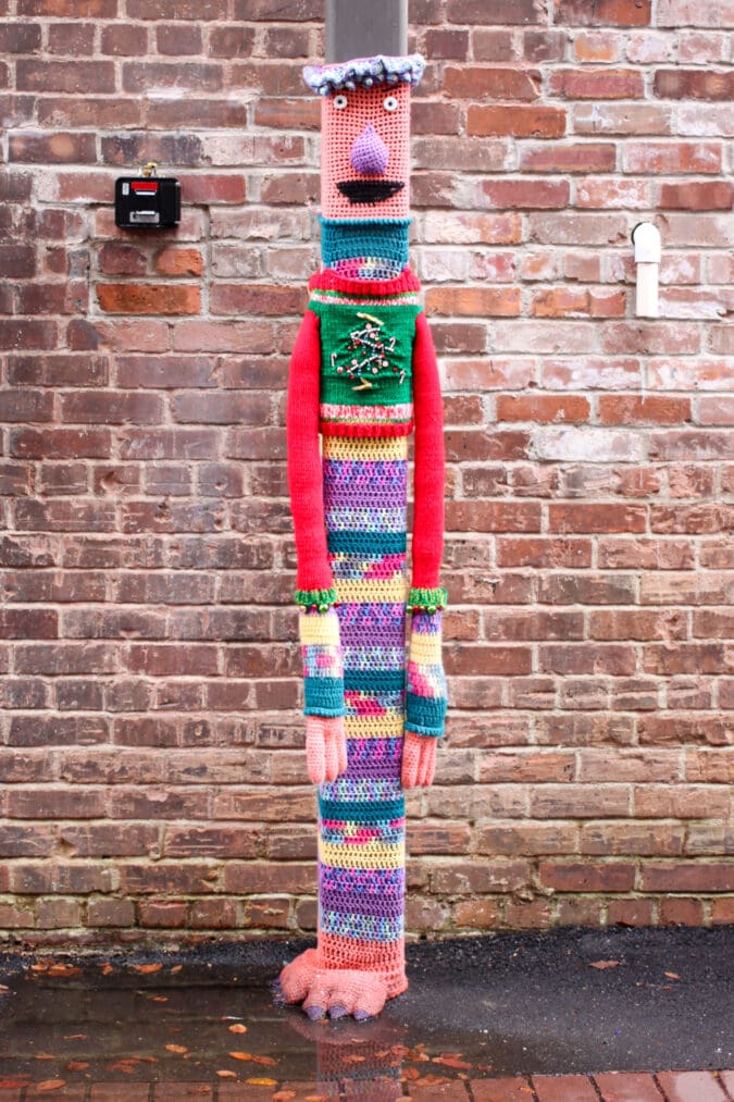 a pole in front of a red brick wall has a colorfully knitted person covering it