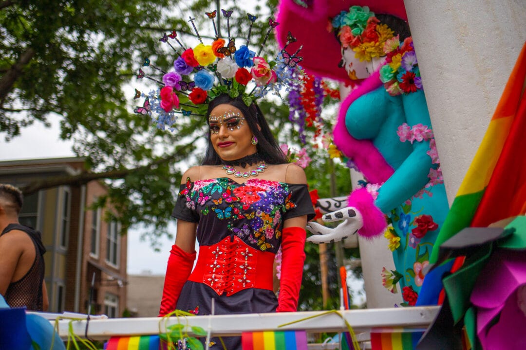 a person wears a flowered headdress and various jewels standing atop a float in a pride parade