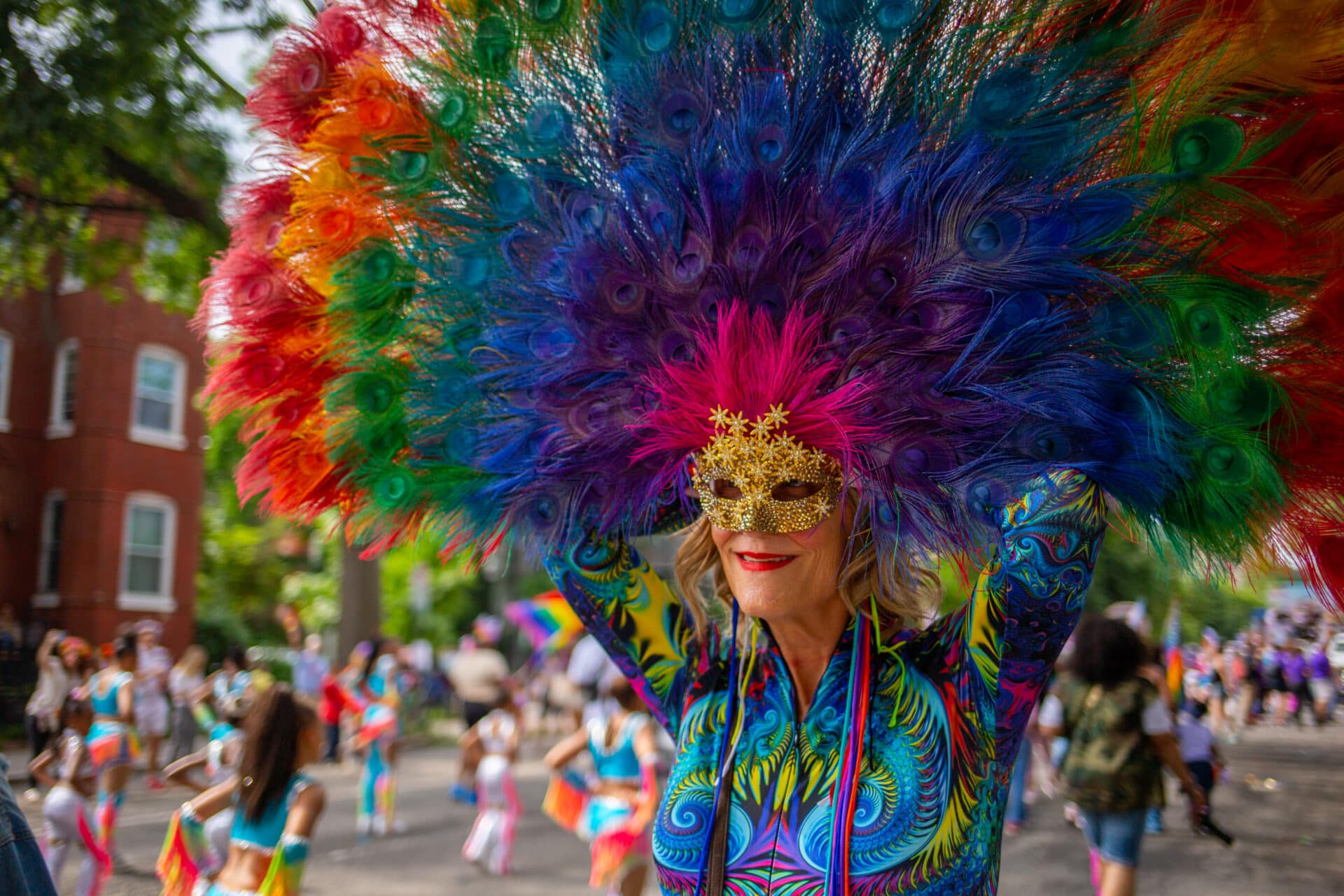 a woman wears a large rainbow peacock feather face mask in a parade