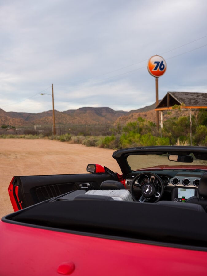 a red convertible is parked outside of an abandoned 76 gas station with the drivers door open