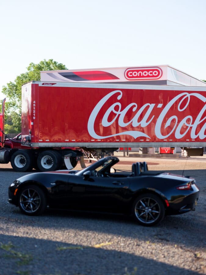 a black convertible is parked next to a semi truck trailer painted with the coca cola logo