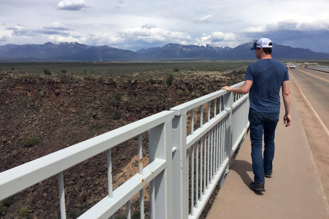 a person walks on a bridge with a white railing over a riverbed with mountains in the background