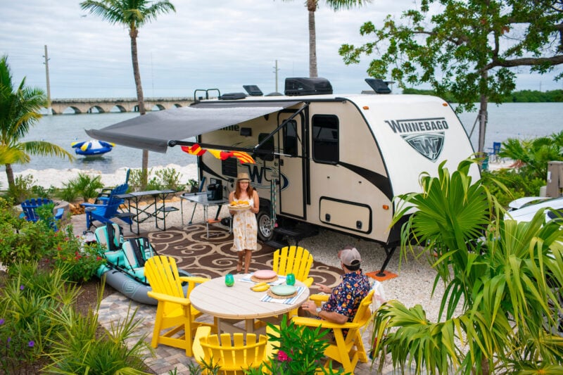 a winnebago is parked at a waterside campsite surrounded by palm trees with a picnic table set up
