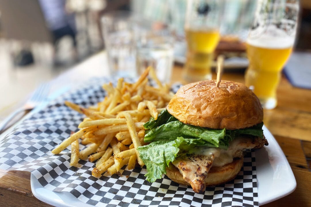 a chicken sandwich and fries on a table with beers in the background