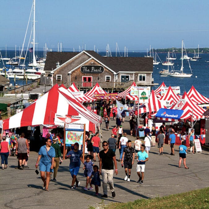 people walk among red and white striped tents near a waterfront harbor full of boats