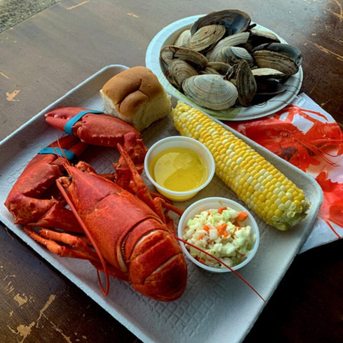 a tray with a whole lobster, corn on the cob, a roll, coleslaw and butter on a table with a bowl of clams