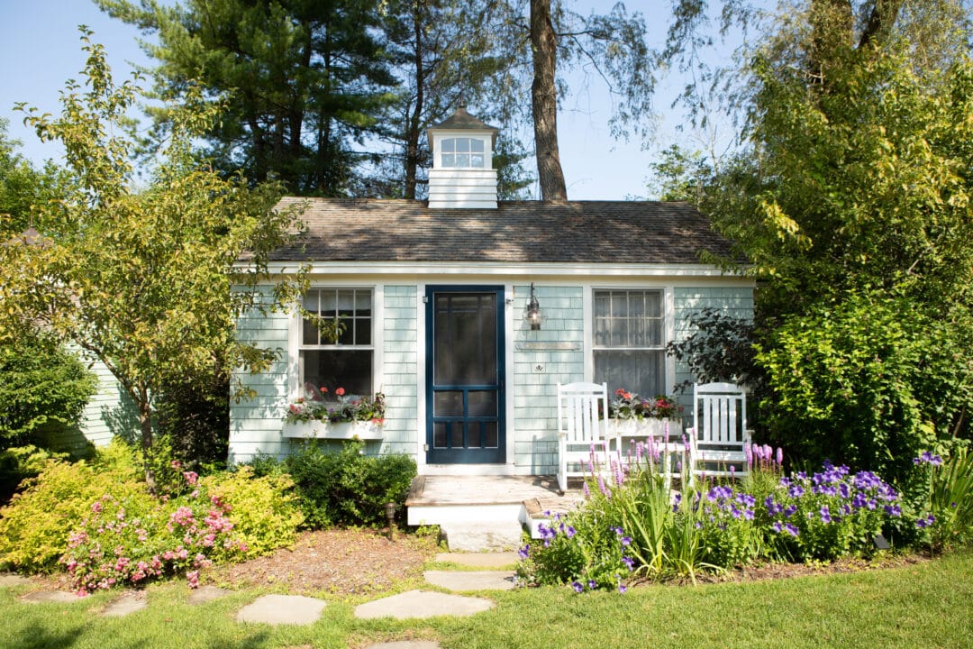 a small cottage with two chairs on the porch surrounded by flowers and greenery