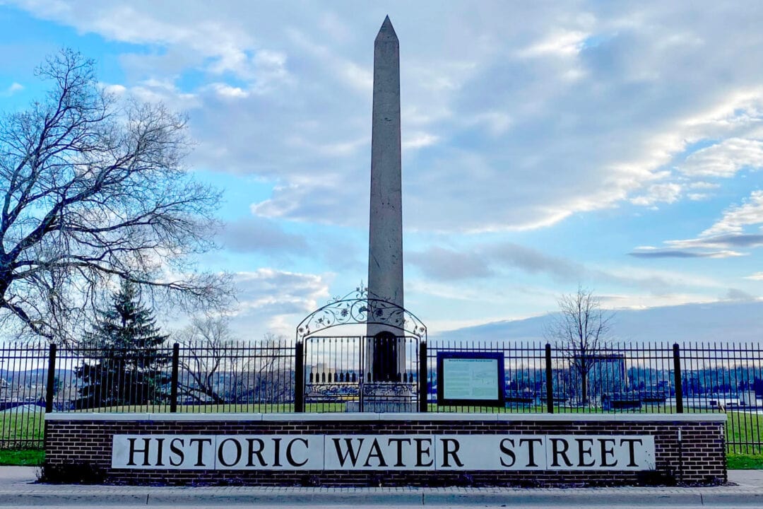 a large obelisk stands behind a black iron gate and fence with a brick sign that says "historic water street"