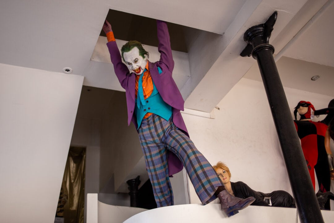a wax figure of a person in clown makeup and a purple suit dangles from the ceiling in a wax museum