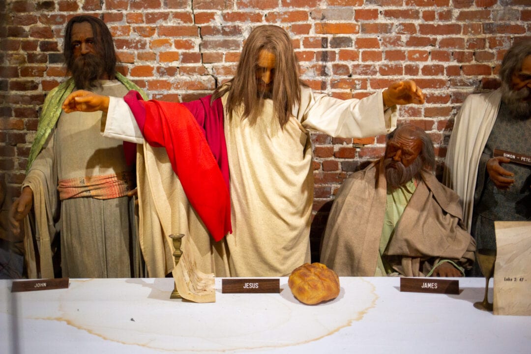 wax figures displayed in the last supper with jesus in the middle with arms outstretched over a while table and a loaf of bread in front of a brick wall