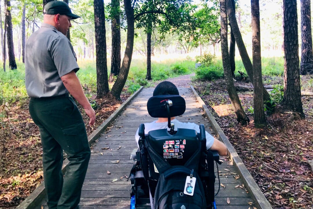a wheelchair user and a park ranger are on a wooden boardwalk in the woods