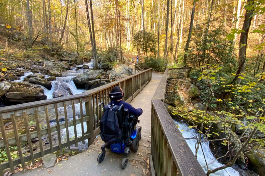 a wheelchair user is on a wooden boardwalk over a waterfall in the woods