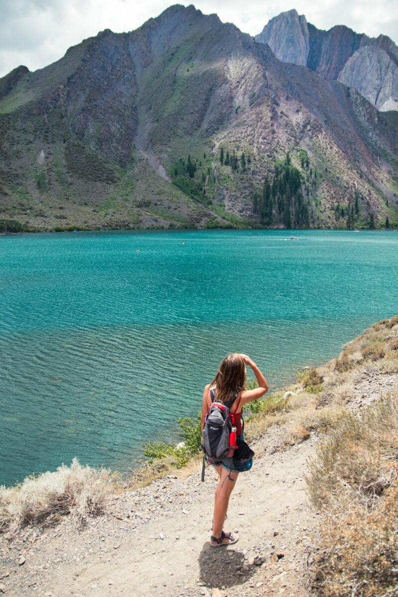 a person stands at the edge of turquoise lake with mountains in the background
