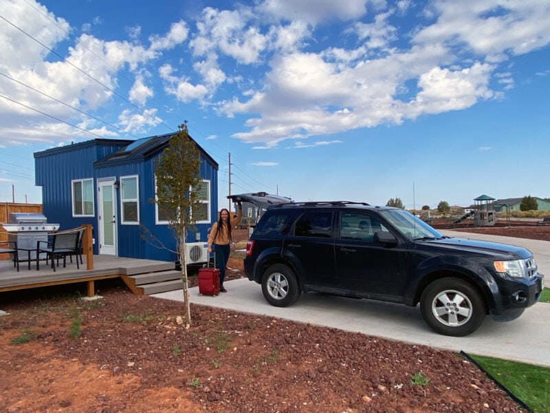 a black suv is parked in front of a small blue cabin with a woman and a red suitcase