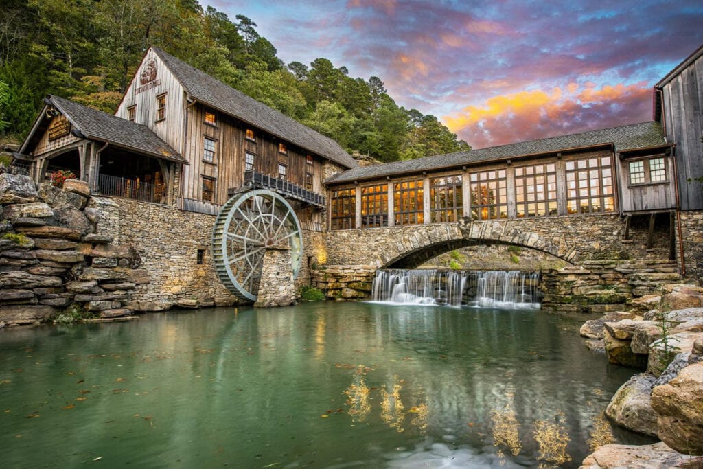 a wooden and stone mill sits atop a rock wall near water at sunset under a pink and purple sky