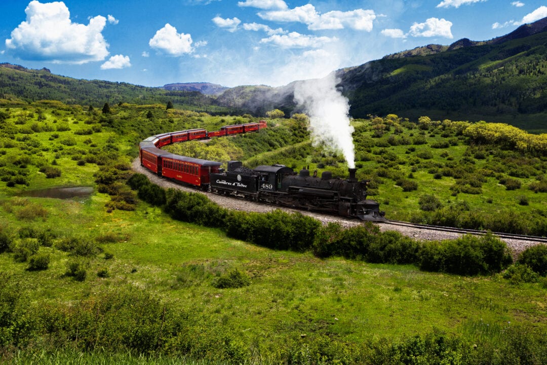 a steam powered train rolls through a green landscape under a blue sky dotted with clouds