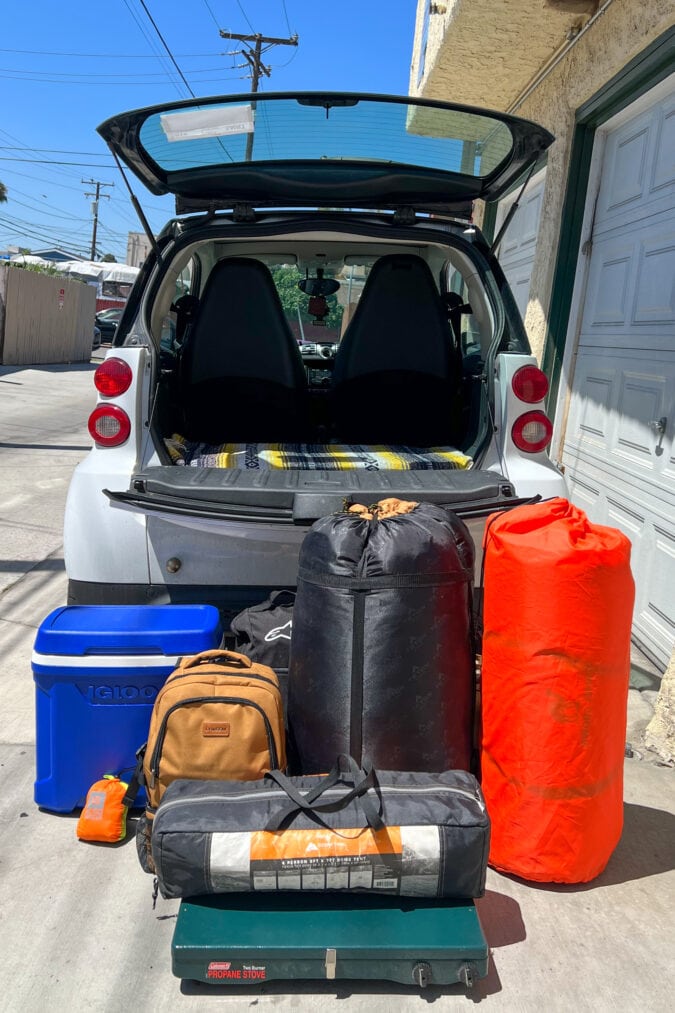 a smart car with it's trunk open and camping gear sitting on the ground