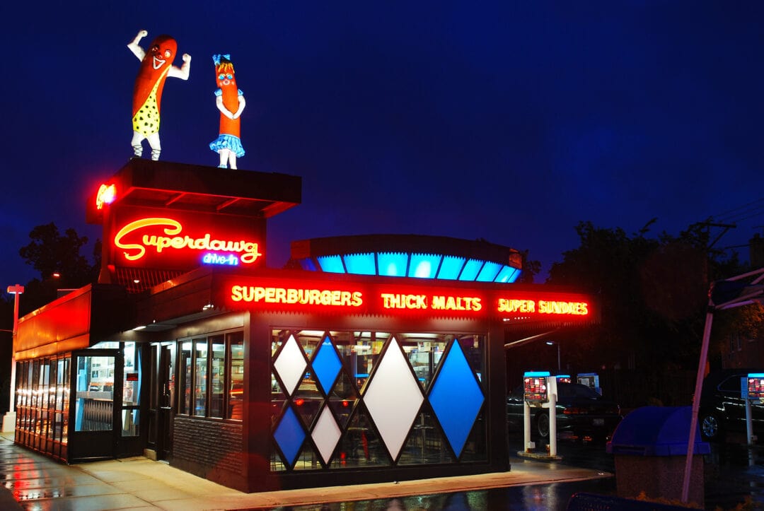 Outside of the Superdawg Drive-In at night with two large illuminated hot dogs standing on top of the building