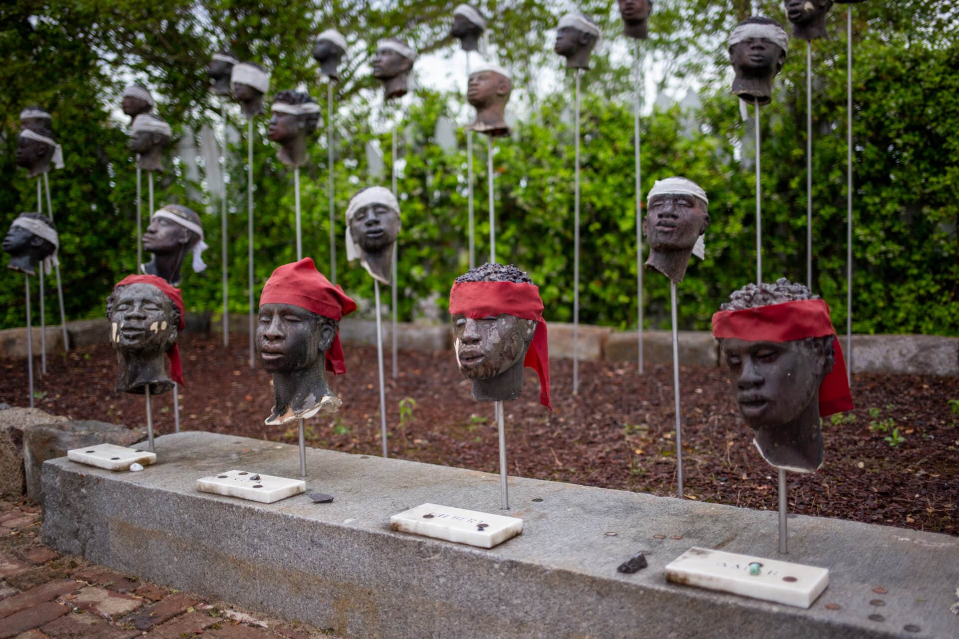 a sculpture depicting several severed heads on sticks, some of which wear red scarves tied around their foreheads, while others have white scarves