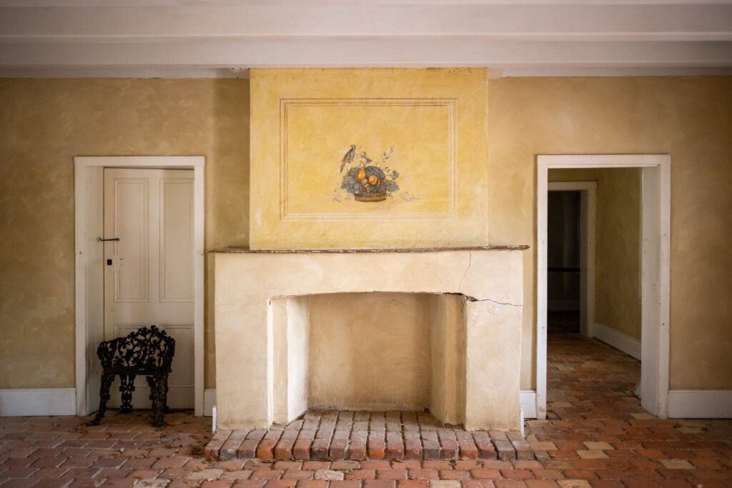 a sparse interior with a chair, fireplace, and painting of fruit above the fireplace