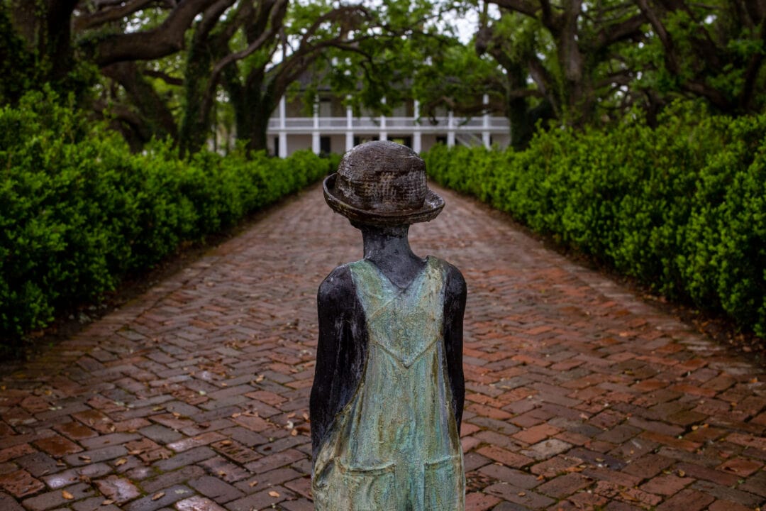 a bronze statue of a small boy in overalls and a hat gazes down a brick path at a large white house surrounded by greenery