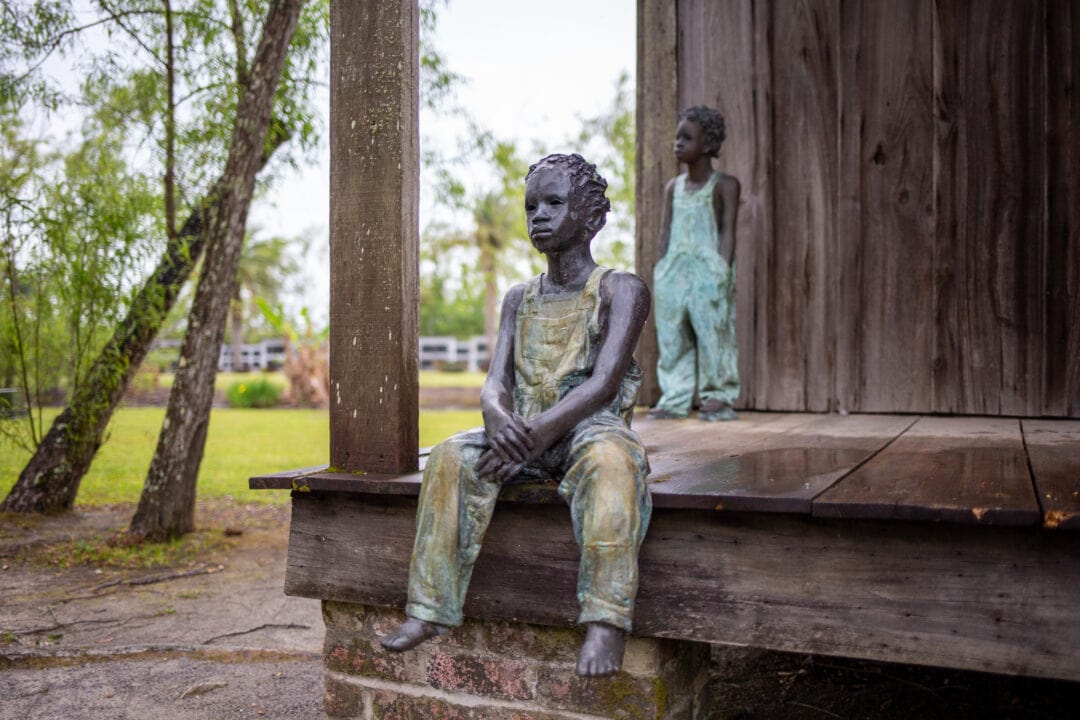 Two sculptures of children sit and stand on the porch of a wooden cabin