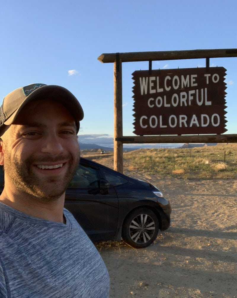 A smiling man in front of a black car and a sign that reads 'Welcome to Colorful Colorado'