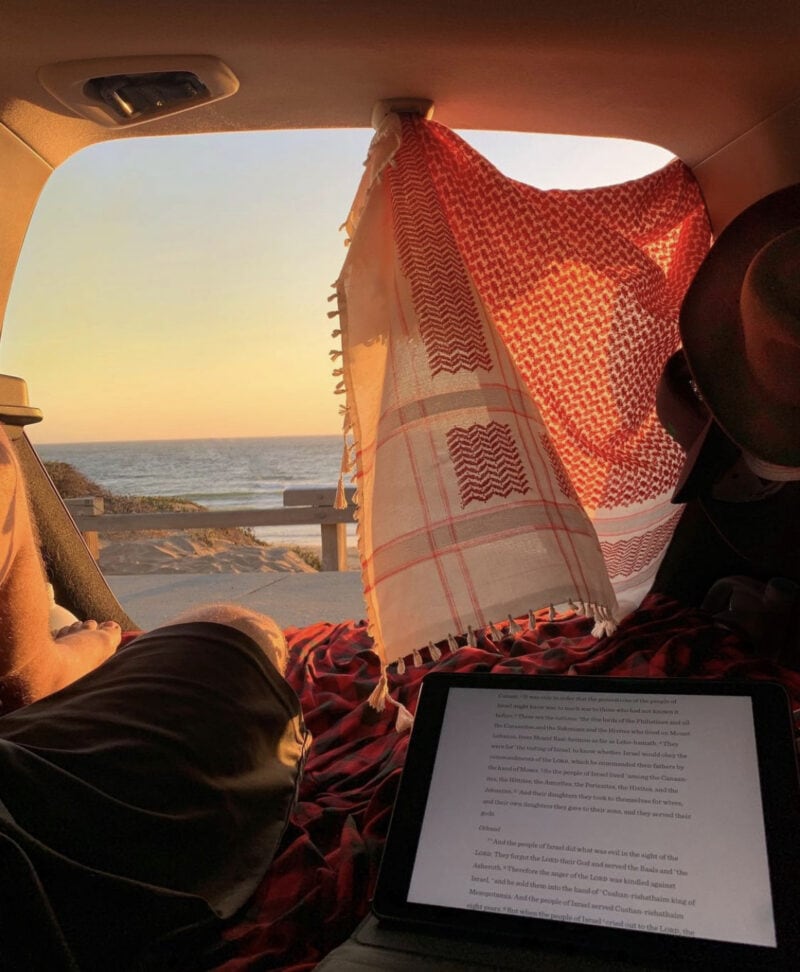 The view of a sunset through an open car hatch is partially blocked by a hanging piece of fabric. An open ebook on a tablet in the foreground.