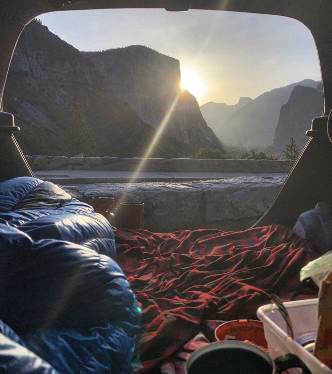 View of a mountain sunset through the open hatch of a car, with blankets and sleeping bags in the foreground