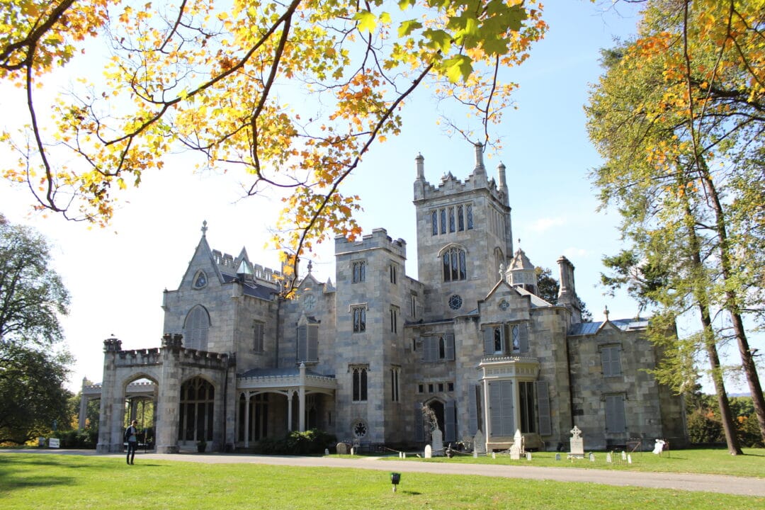 a large grey stone gothic mansion with cardboard tombstones out front surrounded by orange and green fall foliage