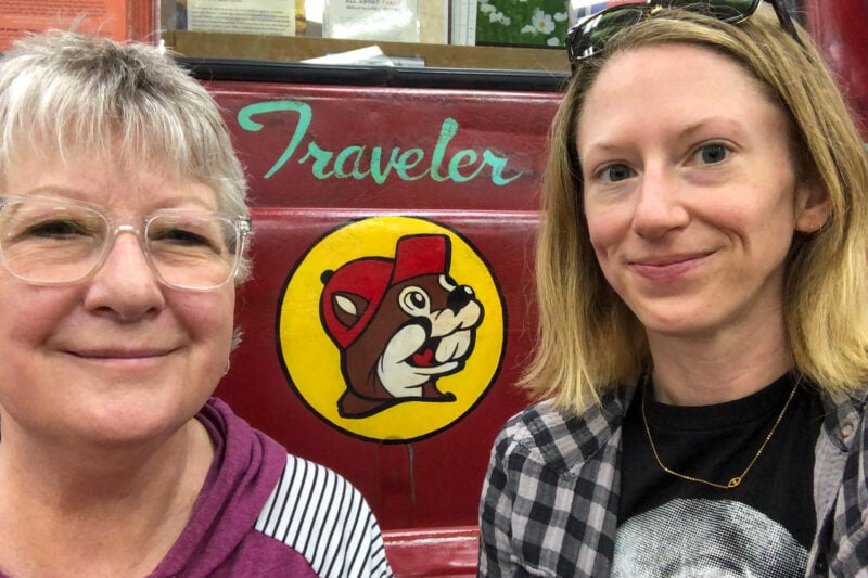 two people take a selfie in front of a red and yellow Buc-ee's beaver logo and the word "traveler"