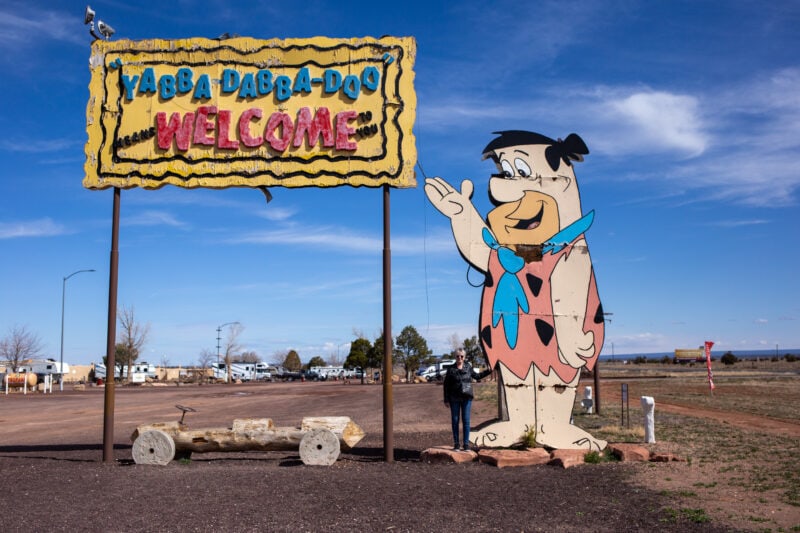 a large wooden sign cutout of fred flinstone with a yellow, blue, and red sign that says "yabba dabba doo welcome"