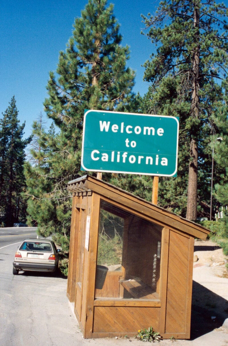 a welcome to california sign on top of a small wooden booth with a car parked out front in front of pine trees