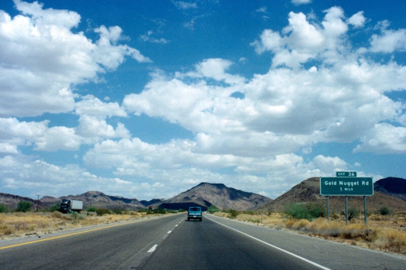 a blue truck driving on a straight road with mountains in the background under a blue sky with fluffy white clouds